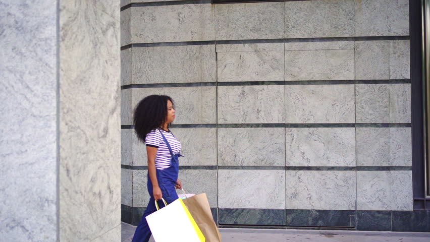 A woman with curly hair walks confidently along a city street holding shopping bags, dressed casually in a striped shirt and denim overalls.