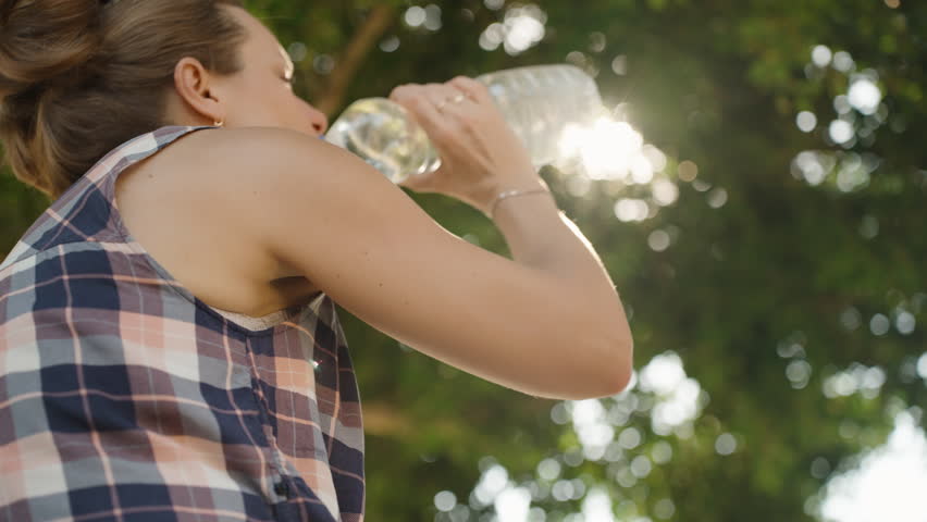 Woman Drinks Water from a Bottle, Hydrating Herself with Bright Sunlight Filtering Through the Trees on a Hot Summer Day.