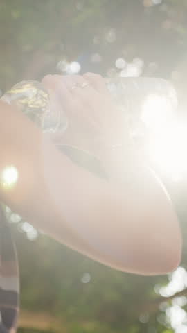 Vertical video. Woman Drinks Water from a Bottle, Hydrating Herself with Bright Sunlight Filtering Through the Trees on a Hot Summer Day.