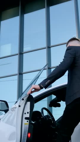 Vertical video businessman in a suit gets into the road in a white car near a glass building in the city. 