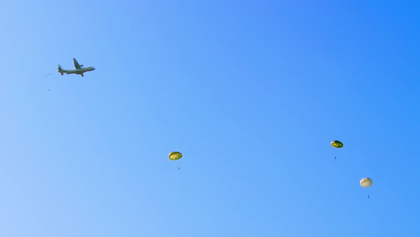 A military aircraft releases paratroopers who descend under round parachutes against a bright blue sky. Captured during a military airborne training exercise at Ginkelse Heide, Netherlands