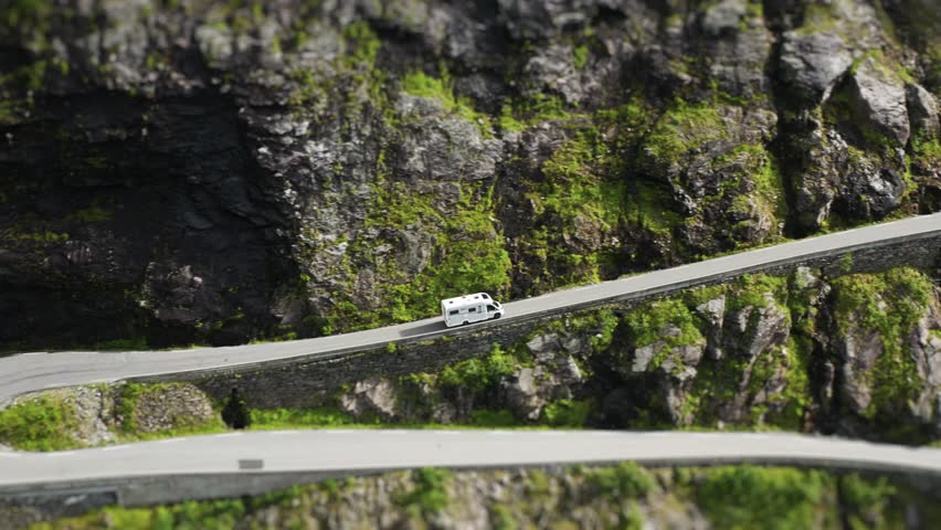 A motorhome climbs a winding Trollstigen road which follows the steep cliffs near a cascading waterfall, offering a breathtaking view. an aerial tilt-shift video.