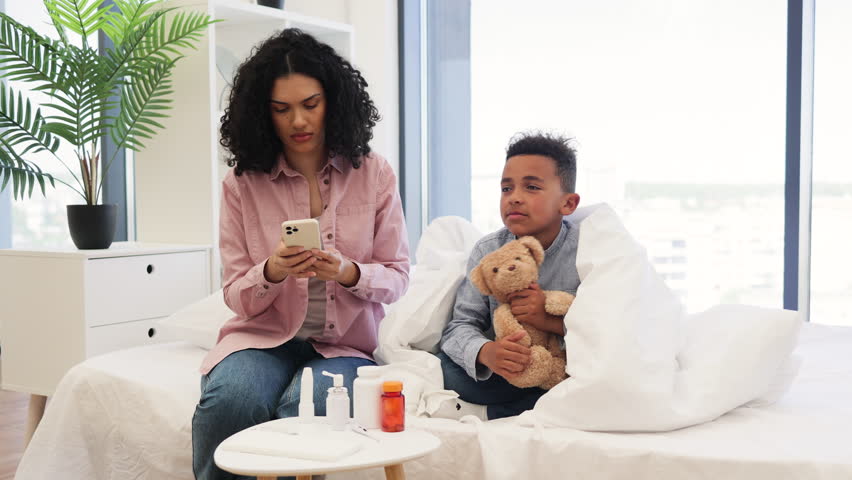 African-American mother checks sick son's temperature while speaking to doctor on phone. Young boy lies on bed wrapped in blanket holding teddy bear, showing concern and care from parent.