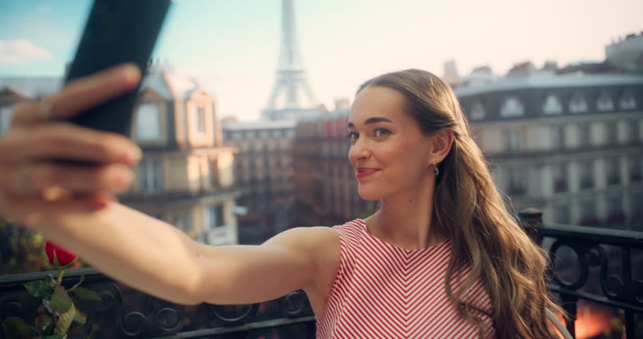 Excited Young Female Captures a Selfie on a Phone on a Parisian Balcony with the Eiffel Tower as a Stunning Backdrop. Her Joyful Expression and Stylish Outfit Adds to the Picturesque Scene