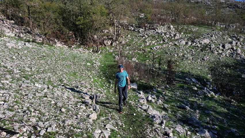 Man in hiking clothes walking towards camera on mountain trail along rocky landscape on sunny day, aerial view