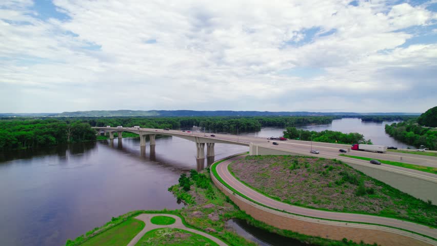 Aerial view of a red semi-truck with a step deck low boy trailer and a dry van trailer crossing the Dresbach Bridge over the Mississippi River at the border of Minnesota and Wisconsin.