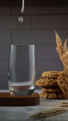 A slow-motion 4K video of milk being poured into a glass on a wooden stand, accompanied by American cookies and dried flowers, creating a cozy atmosphere.