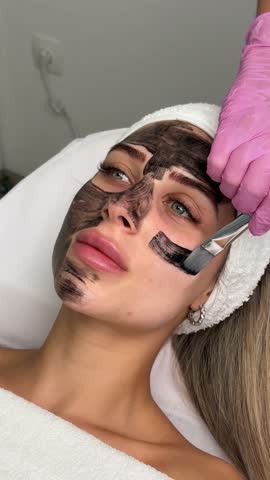 cosmetologist applying black mask on pretty woman face wearing black gloves, gorgeous woman in spa having facial procedures