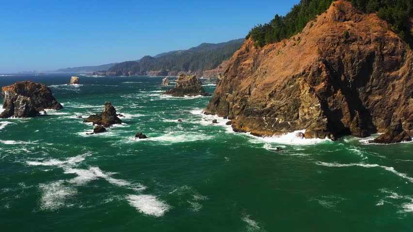 Scenic aerial shot gliding over ocean waves and sea stack rock formations. Shot by an Evergreen forest and cliffside on the shores of Oregon, Pacific Northwest.