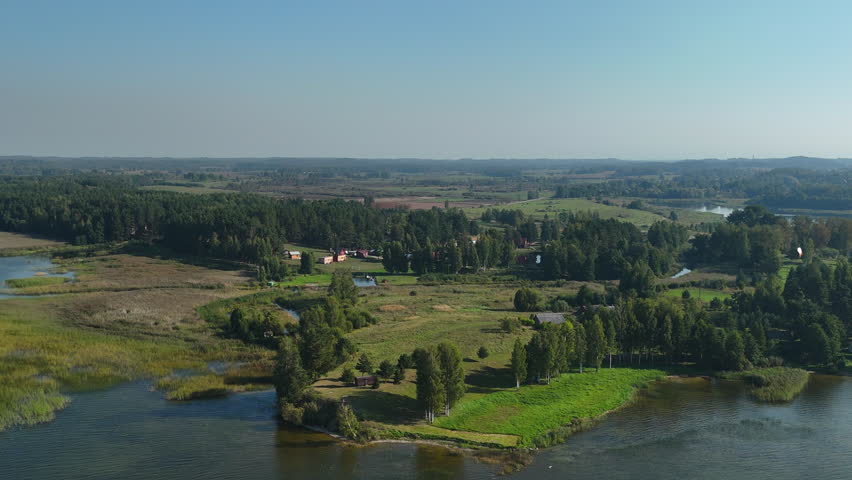 Smooth aerial approach over a peaceful lakeside village surrounded by lush greenery and forests in Latvia. Ideal for nature, rural landscapes, and travel footage, showcasing serenity and natural beaut