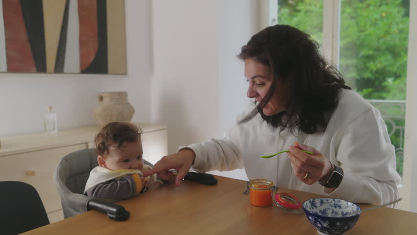 Mother feeding baby while sharing an affectionate moment, baby smiling and looking up at mother, playful interaction during mealtime, mother holding spoon with baby food
