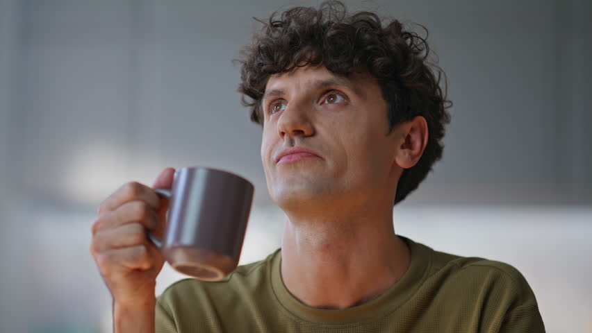 Dreamy man drinking coffee pondering project at home workplace closeup. Smiling remote employee sitting at living room. Happy curly entrepreneur thinking on business success at modern domestic office