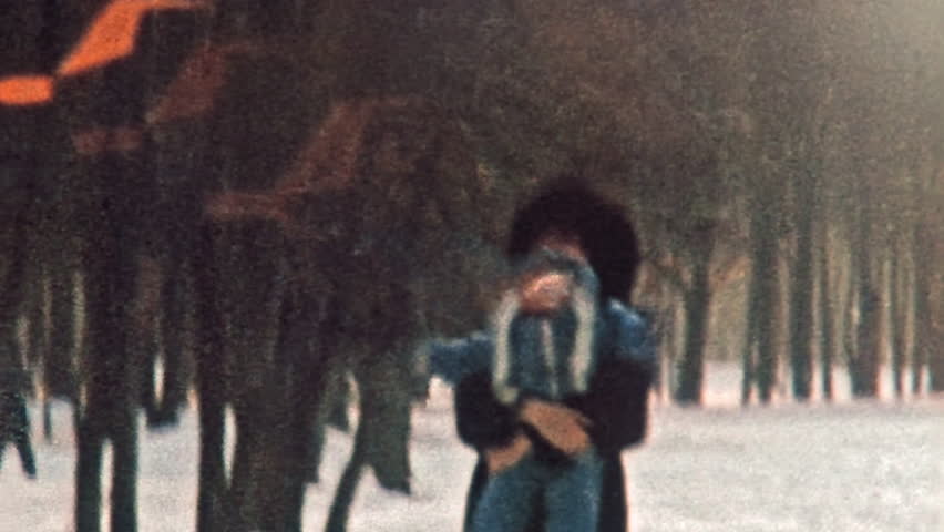Happy mother holding baby in winter park outdoors. Smiling toddler running through winter snow towards camera in sunlight. Happy family vacation in cold sunny nature. Vintage film. Retro 1980s archive