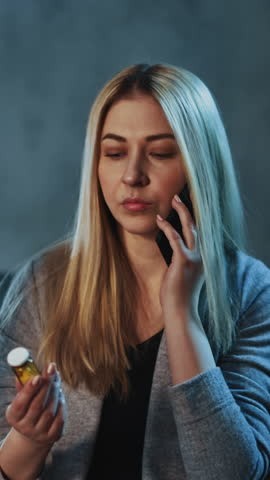Doubting blonde woman calls on smartphone holding small pill bottle against workplace with glowing lamp in semi-dark room close view slow motion