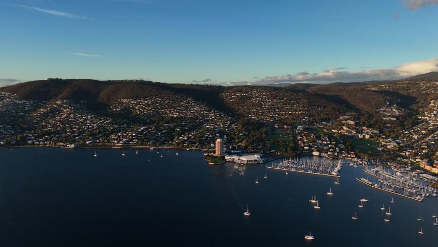 Beautiful coastline of Tasmania with city of Hobart during golden sunset. Aerial panorama panning shot. Luxury marina and port with housing area and neighborhood on hillside.