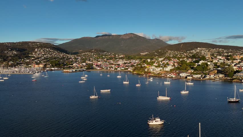 Drone approaching shot of anchored boats and yachts at waterfront of Hobart City. Sunset time with mountain range in background. Buildings and homes located on hillside.Wide shot.