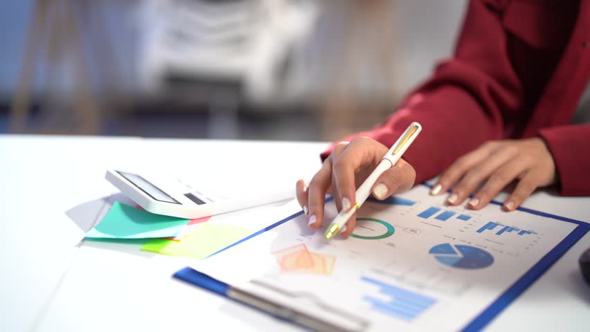 Accountant woman analyzing financial data, pointing at charts and using a calculator in an office setting. Focus on her professional approach to examining graphs and reports
