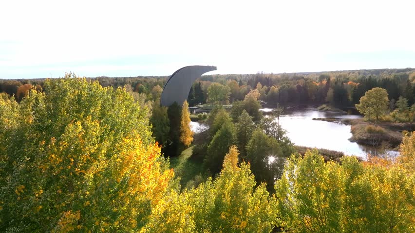 A modern curved observation tower stands amidst a vibrant autumn landscape, surrounded by golden and green trees, dense forest, and open fields.