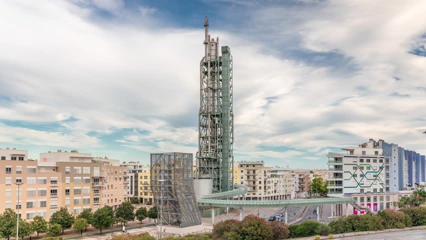 Timelapse hyperlapse of old steel refinery tower with spiral ramp in Lisbon's Park of Nations modern district. Traffic and cloudy sky in the background. Apartment buildings around. Portugal
