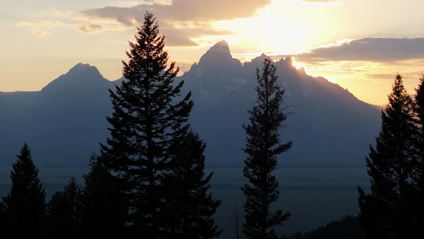 Grand Tetons at sunset with tree silhouettes, warm orange glow fading behind mountain peaks