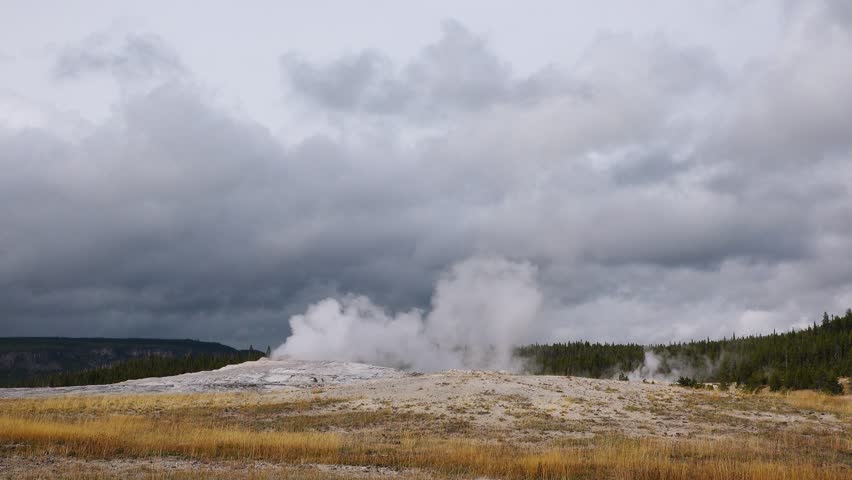 Spectacular eruption of Old Faithful Geyser in Yellowstone National Park, as steam and water erupt dramatically into the air, against a backdrop of dark, swirling clouds.