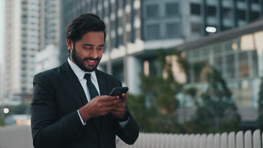 Unshaved businessman in black suit looking at screen of cellular phone with excited smile while standing close to modern buildings. Carefree person texting online while using application with internet