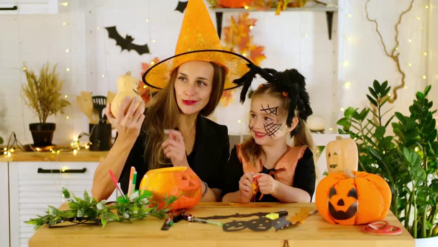 Halloween, a happy family a mother with a baby girl in festive costumes and makeup is preparing for Halloween at home in the kitchen by coloring and carving pumpkins and scaring everyone