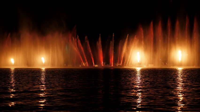 A beautiful fire show and a show of colorful fountains on a small lake. Flashes of fire and jets of water in the dark.