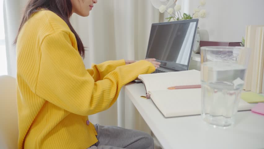 Asian girl student drink water while learning online class study with teacher in house. Teenage woman smiling, taking notes and using laptop computer device remote study on table in bedroom at home.