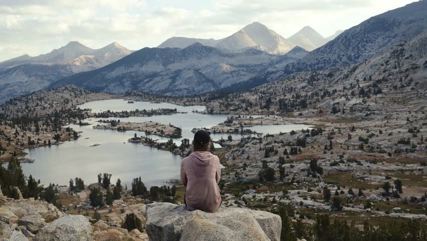 Hiker sits on a rock overlooking scenic lakes and mountain peaks along the John Muir Trail