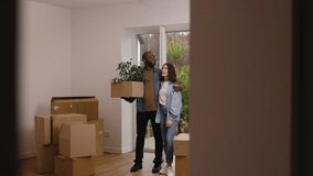 A young couple is moving into their new home. The man, holding a box with a houseplant, kisses his partner on the forehead as she lovingly embraces him. Several moving boxes are visible in the room - Powered by Shutterstock - Get 15% off with code: PIKWIZARD15
