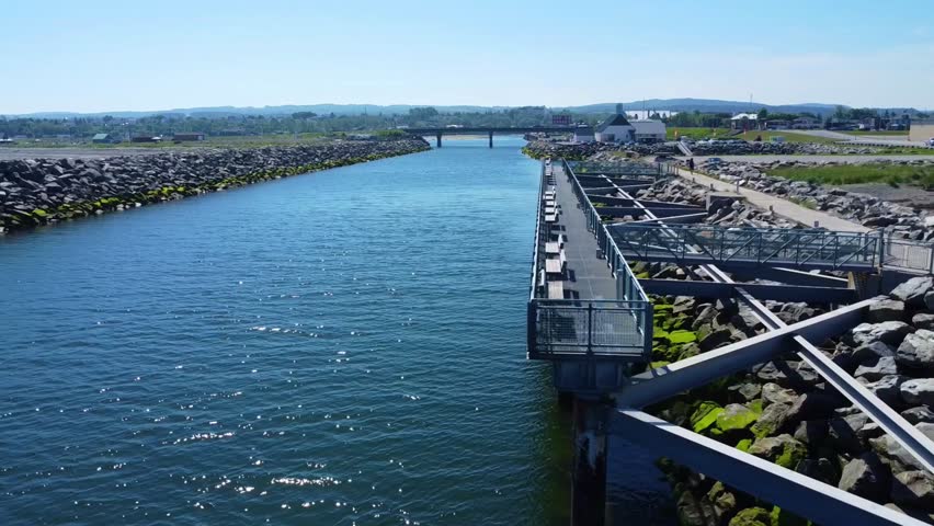 Metal structure footbridge for fishing and observation built along an artificial rock channel at the mouth of the Matane River leading to the Road 132 bridge under a blue sky. Gaspe penisula, Quebec.