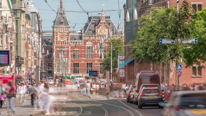 Damrak Street heading from Dam Square to Centraal Station in a timelapse with electric tram. Traffic moves on the road while people wait at the tram stop. Amsterdam, Netherlands