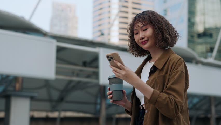 Beautiful Young Woman Walking Through The City Holding Phone And Coffee. Pretty Asian Woman Surfing Internet During Coffee Break. People and Technology Concept