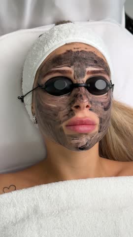 Close-up of cosmetologist's hand making hardware carbon peeling by young pretty customer woman in safety glasses headdress lying on couch in beauty salon. Facial cleaning concept. Cosmetology Services