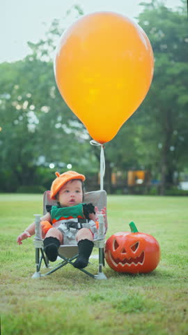 Cute small baby girl sitting near pumpkin with orange balloon in park outdoor, Thanksgiving and Halloween. Festive season in October.