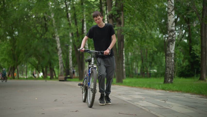 White young boy in black top is walking his bicycle along a peaceful park pathway, surrounded by lush green trees and nature, people can be seen walking in the softly blurred background