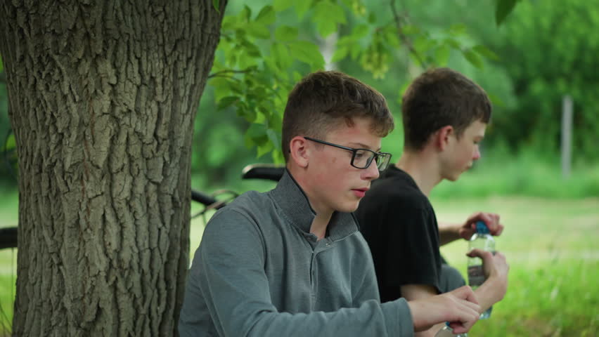 Two young siblings squat under a tree, drinking from water bottles while resting after a bike ride, the bicycles are parked nearby, with a blur view of someone riding pass by with a bicycle