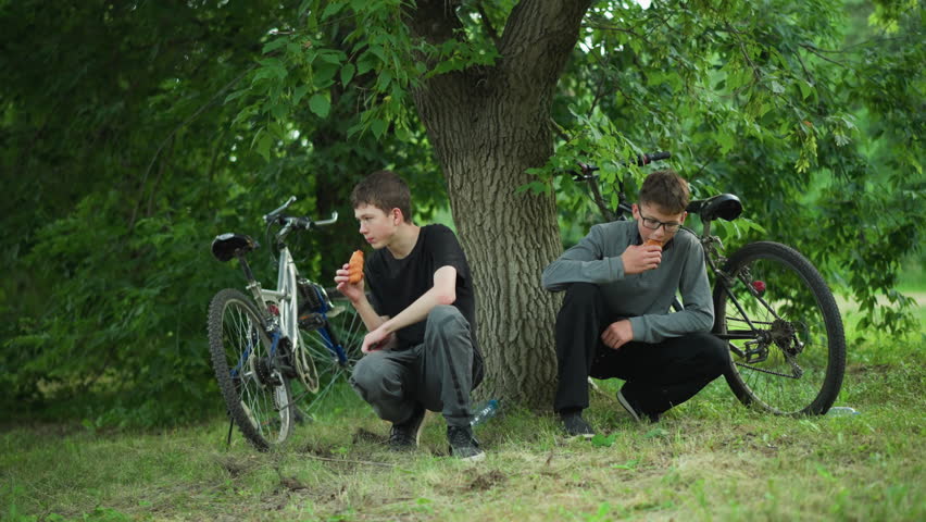 Two young siblings enjoying snacks while squatting under a tree in a lush green forest, they are taking a break from cycling, with their bikes parked close by