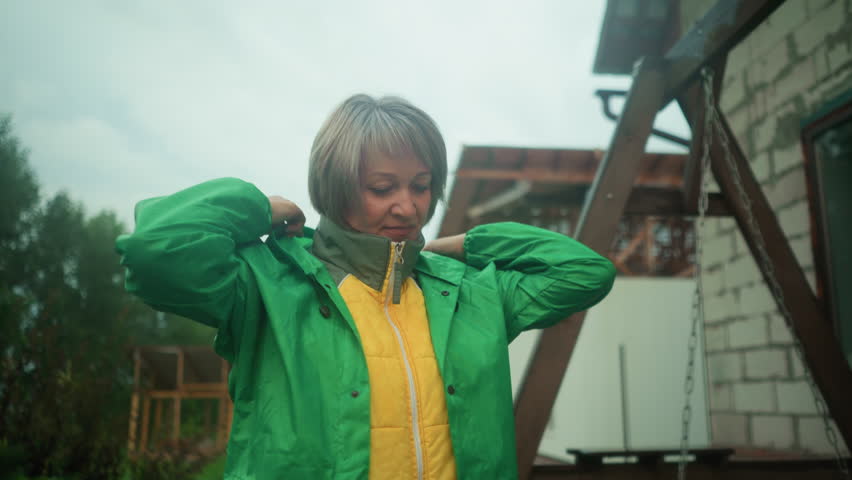 Middle-aged woman wearing yellow jacket under green raincoat adjusts hood while standing near swing bench, background features blurred greenery and building