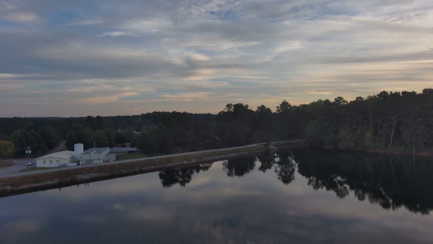 aerial footage of the of the still blue lake in the park at sunset surrounded by lush green trees with blue sky and powerful clouds at Rhodes Jordan Park in Lawrenceville Georgia USA