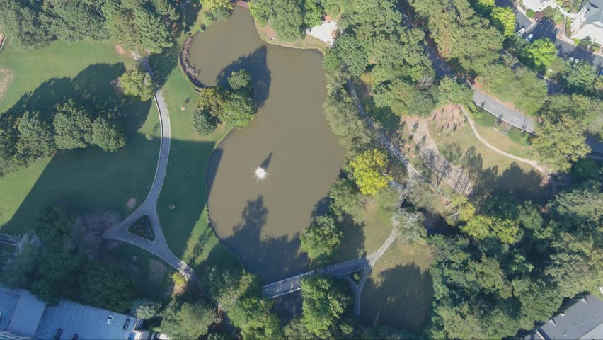 aerial footage of a silky green lake in the park with a water fountain surrounded by lush green trees and grass at Lenox Park in Brookhaven Georgia USA