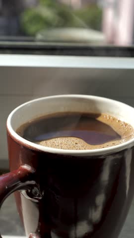Vertical: A brown cup of hot coffee with foam releases steam upwards standing by the window. Morning invigorating coffee drink for waking up. Freshly brewed ground coffee in a mug. Espresso, Americano