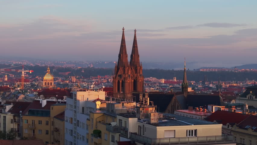 Sunlight washes over the Prague skyline, casting long shadows from the Bazilika sv. Ludmily and other landmarks at sunset