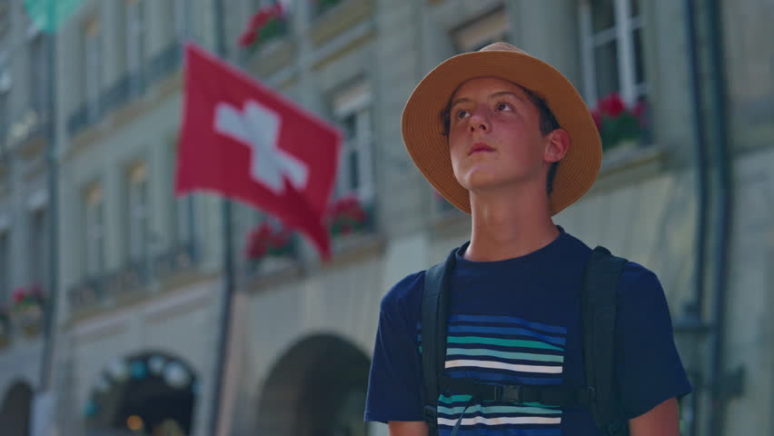 Young guy student tourist with backpack and hat in the center of the old city of Bern watching architecture, buildings around on the background of the Swiss flag. Switzerland