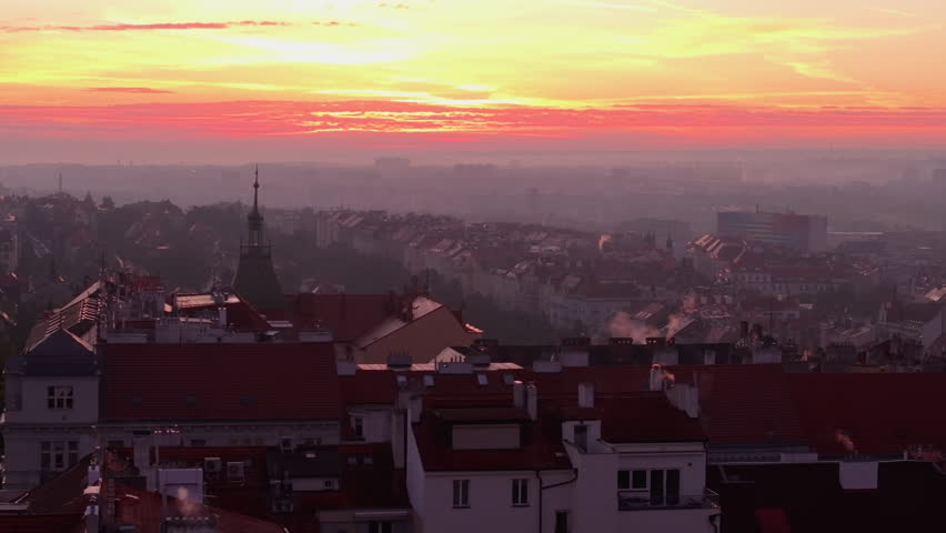 Orange, yellow and purple colors fill the sky as the sun sets above Prague. Smoke rises from chimneys as the city waits for the night to come