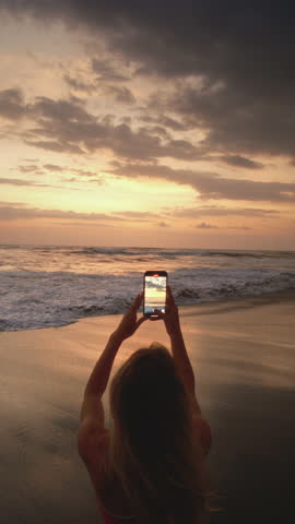 Vertical: Sunset, phone and beauty sexy woman taking a photo while traveling on the ocean or sea in tropical country. Smartphone, female tourist on vacation trip taking a picture at Bali twilight