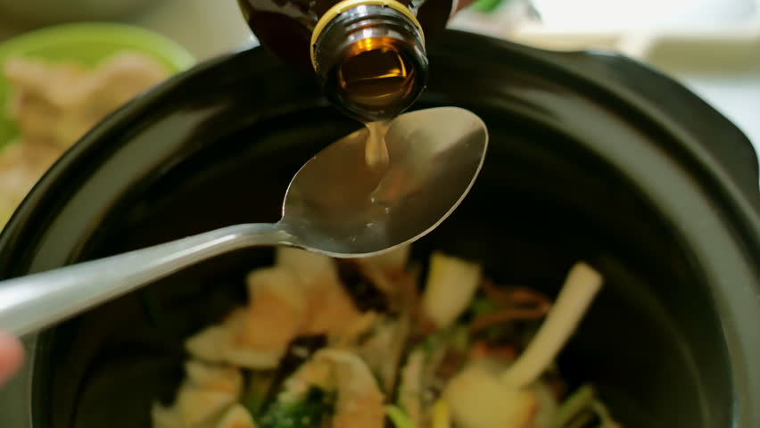A closeup shot captures a spoonful of apple cider vinegar being poured evenly into a black pot of soup filled with dry bouillon, star anise, onion, green onions, celery, carrots, and pandan leaves