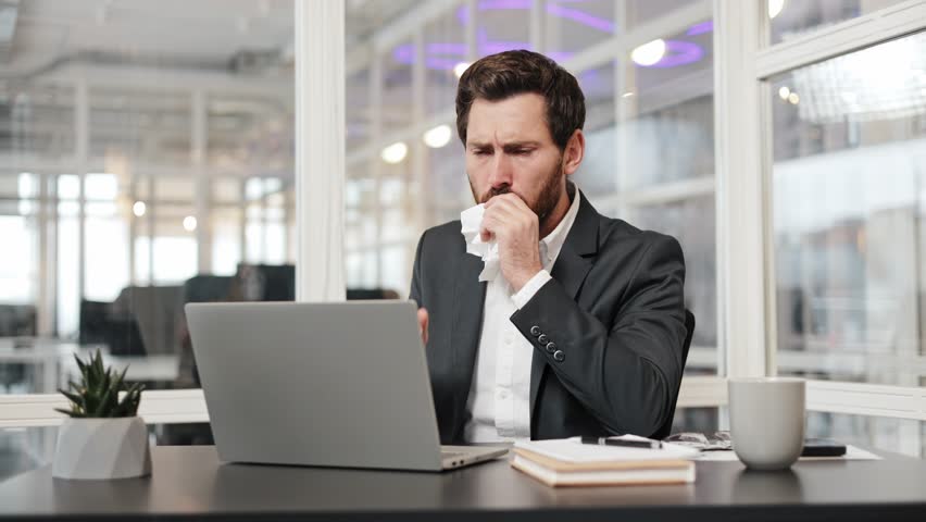 Ill businessman in a suit sits at his desk and coughs into a tissue. Banking worker is sick and working from the office. Man has a laptop, and is looking concerned.