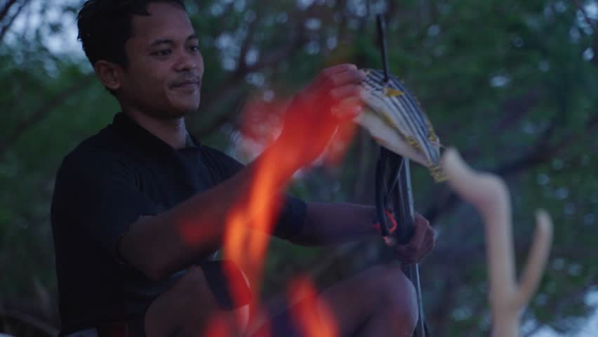 A happy spear fisherman taking two fish that he has caught off his speargun with a fire burning in front of him ready to cook his fish